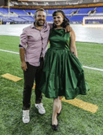 Male wearing black pants and shirt and female wearing green dress standing on a sports arena field 